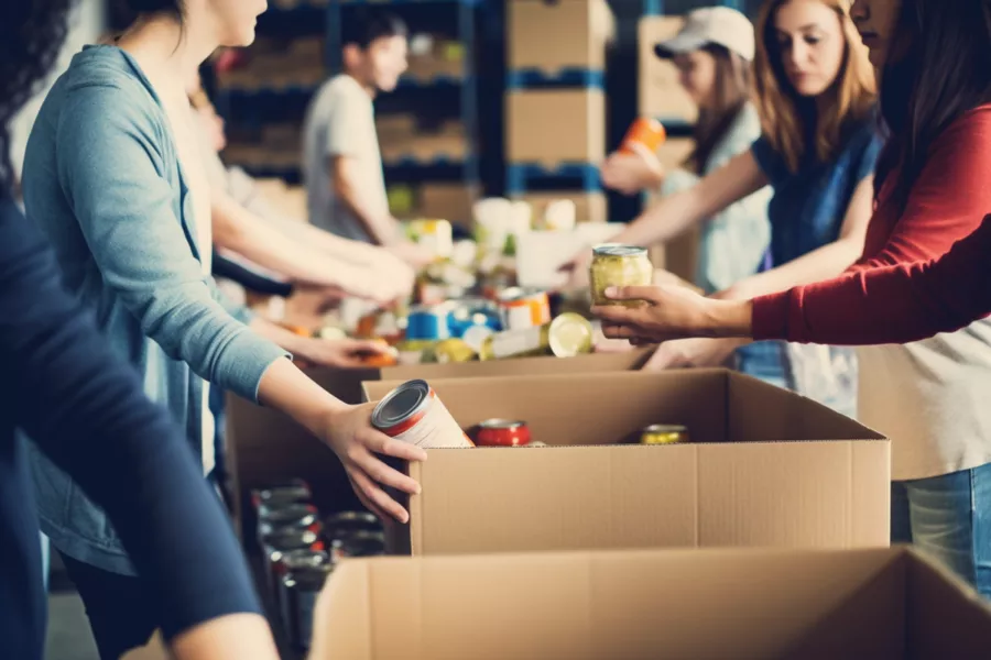 A group of people pack canned goods and jars into large cardboard boxes on a table, likely volunteering at a food bank or donation center.
