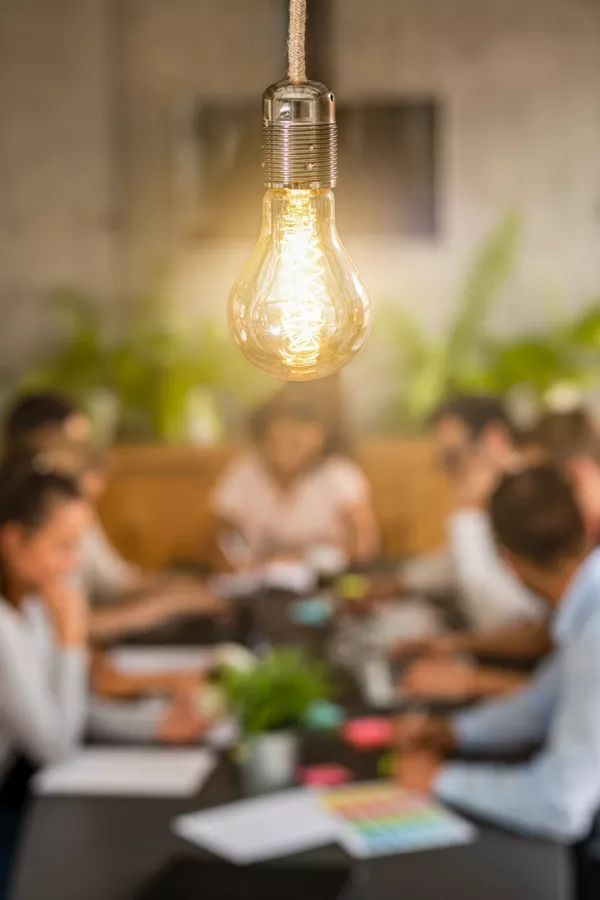 Close-up of a lit hanging light bulb with a group of people working together at a table in the blurred background.