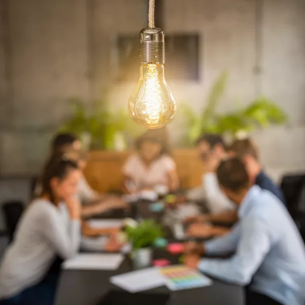 A lit light bulb hangs in focus above a table where a group of people are having a meeting, with plants and office materials in the background.