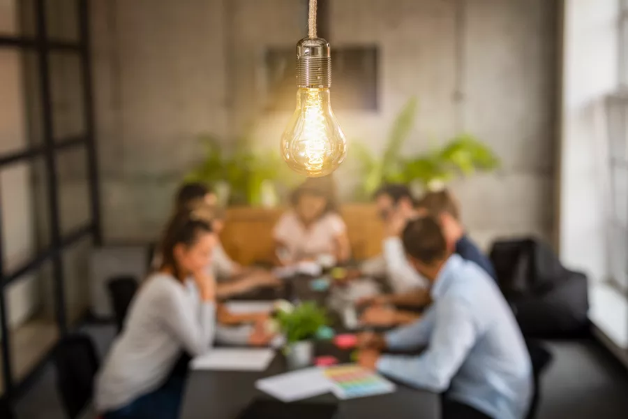 A lit light bulb hangs in focus above a conference table where a group of people are having a meeting in a modern office setting.