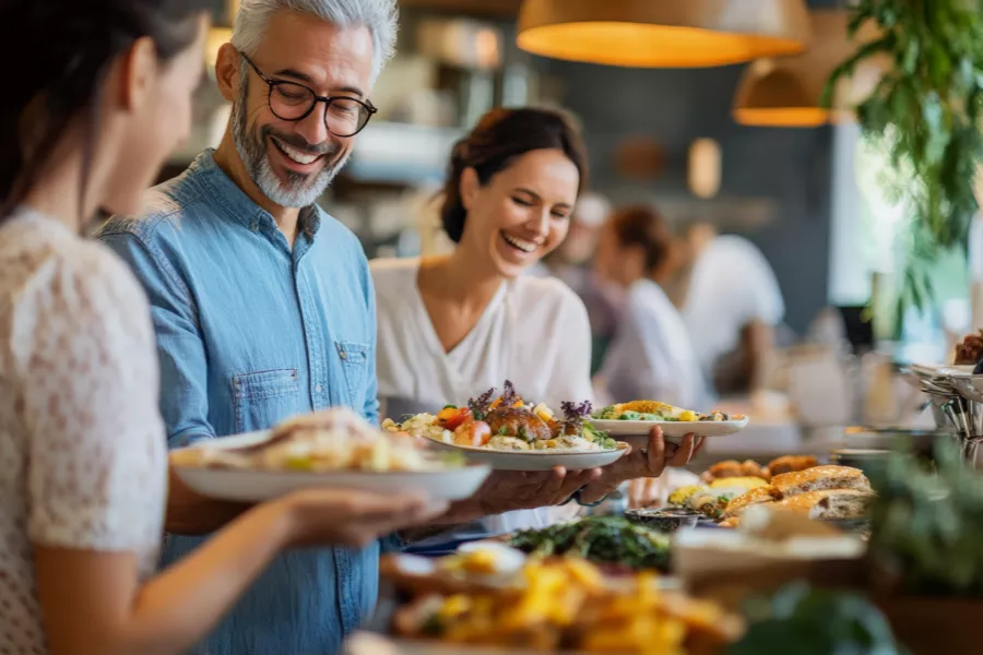Three people stand in a restaurant holding plates of food, smiling and talking, with a buffet spread and other diners visible in the background.