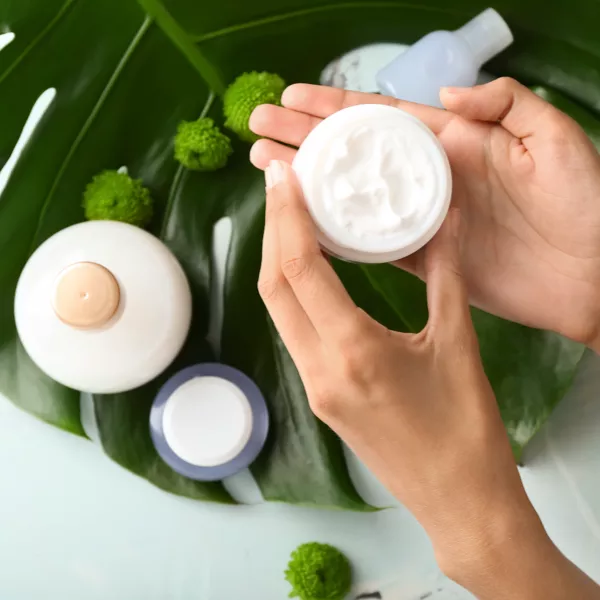 Hands holding an open jar of white cream above a large green leaf, with two closed bottles and some small green flowers nearby.