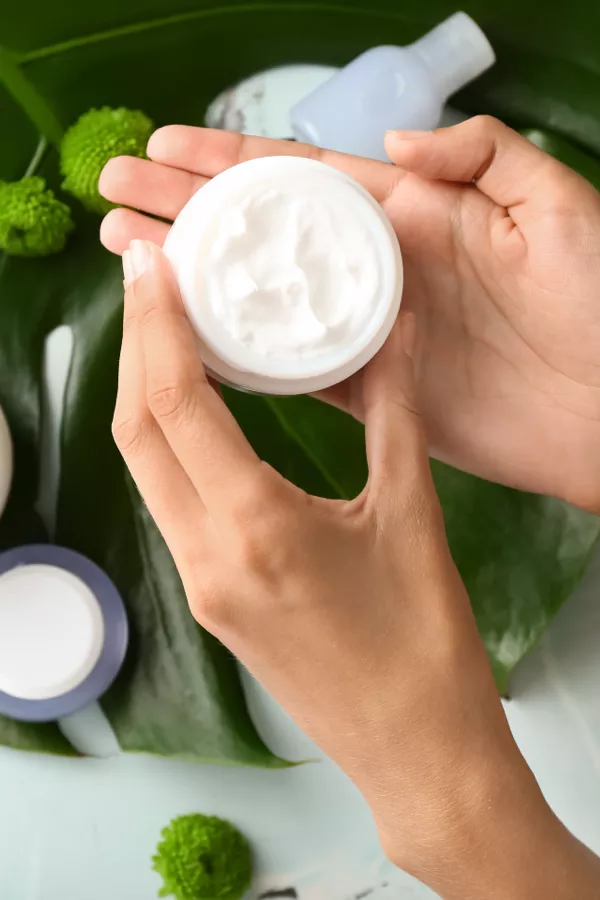 A person holds an open jar of white cream above green leaves, with other skincare containers and decorative green spheres nearby.
