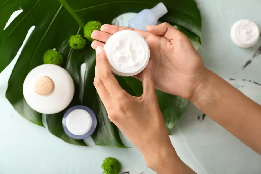 Hands holding an open jar of cream above a large green leaf, with two closed bottles and decorative green accents nearby.
