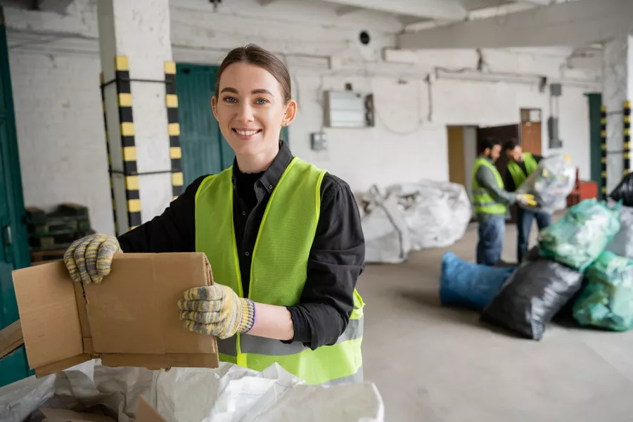A woman wearing a high-visibility vest and gloves smiles while handling cardboard in a recycling facility, with coworkers sorting materials in the background.
