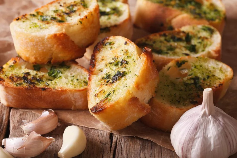 Slices of toasted garlic bread with herbs are arranged on parchment paper, accompanied by whole and peeled garlic cloves on a rustic wooden surface.