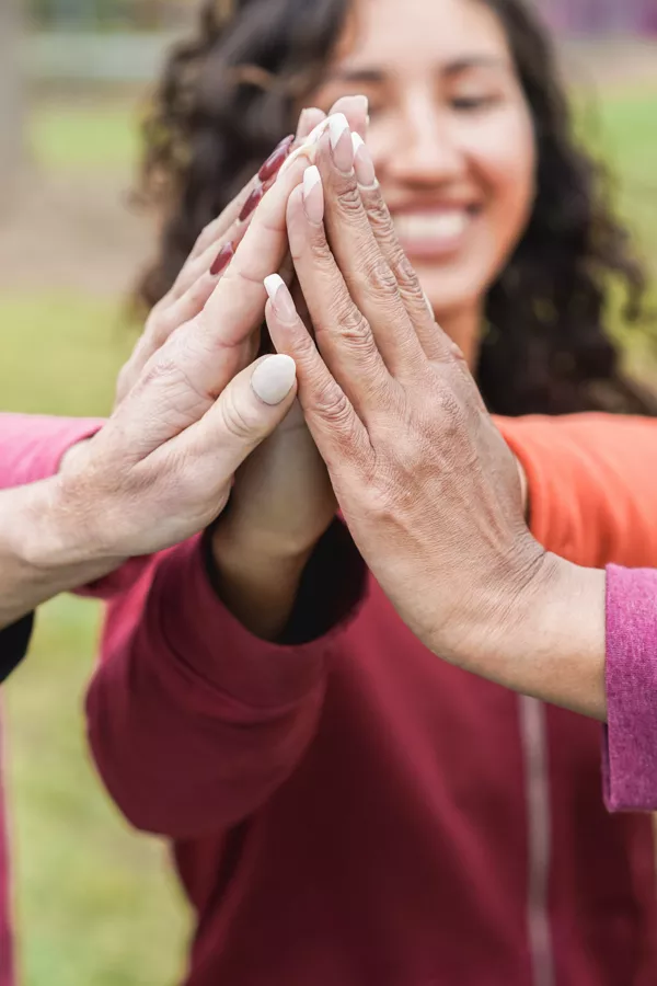 Three people join hands in a group high five outdoors, with a smiling person in the background wearing a red jacket.