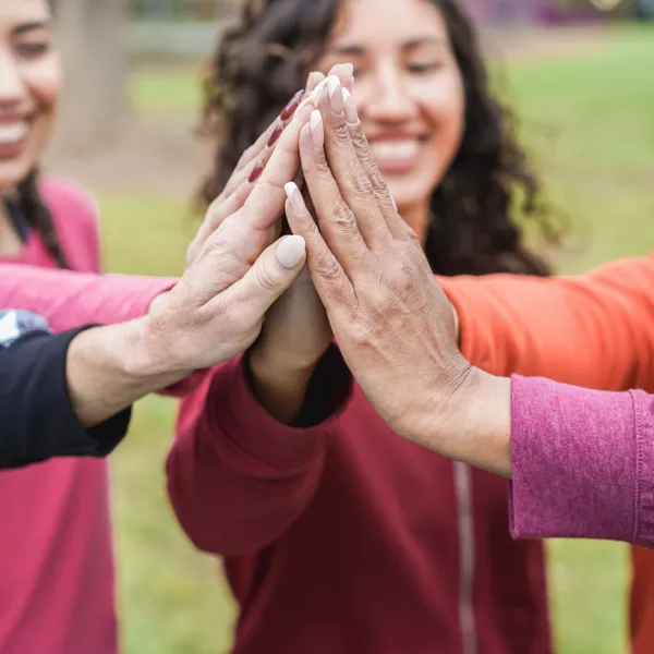 Three people standing outdoors smile as they bring their hands together in the center for a group high-five.