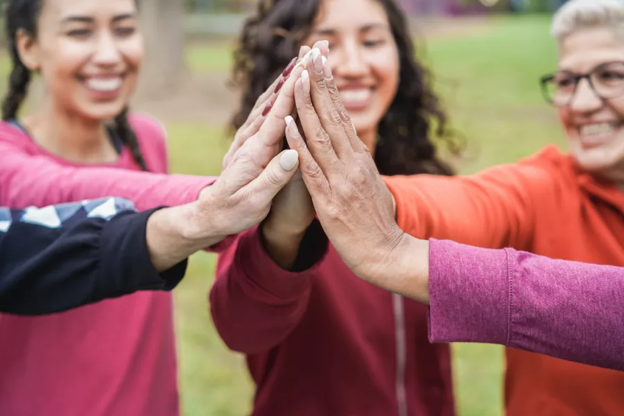 Four people standing outdoors in a circle give a group high five, their hands meeting at the center, with smiling faces visible in the background.