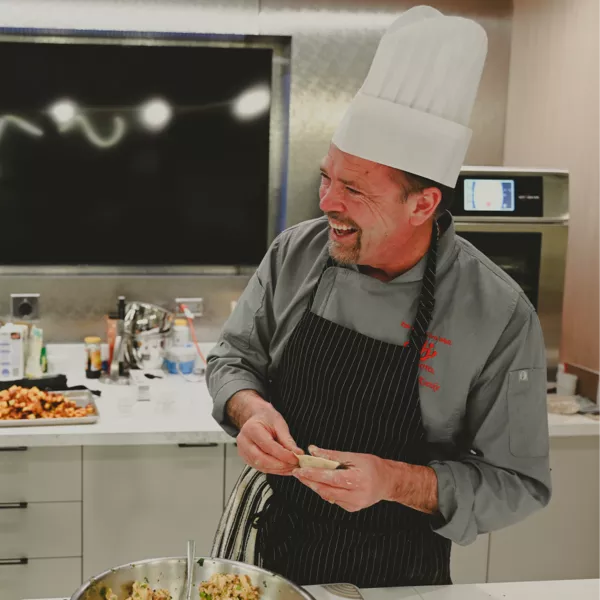 A chef wearing a white hat and striped apron smiles while preparing food in a modern kitchen.