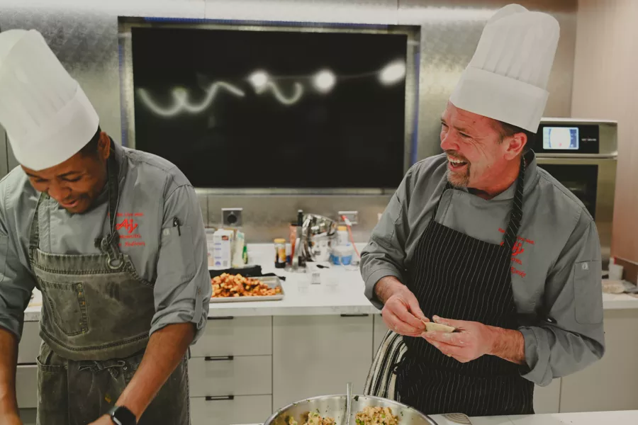 Two chefs in uniforms and white hats prepare food and smile together in a modern kitchen.