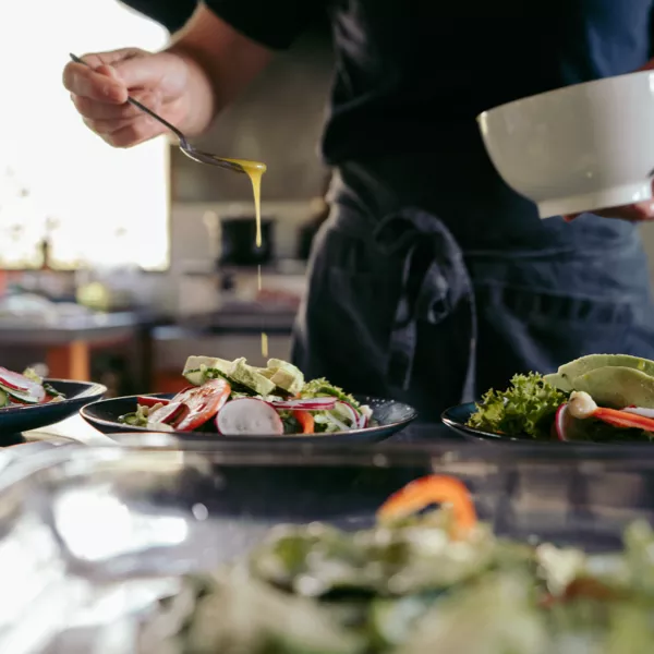 A person drizzles dressing over a salad with a spoon while holding a bowl, with plates of salad arranged on a kitchen counter.