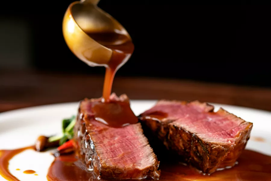 A close-up of two slices of rare beef steak on a plate, with brown sauce being poured over them from a ladle. Vegetables are visible in the background.