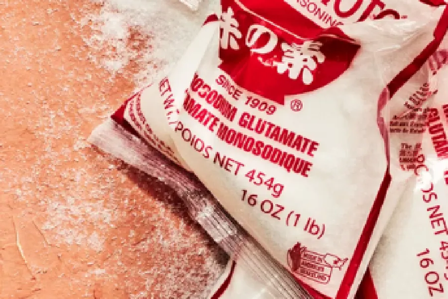 A bag of monosodium glutamate seasoning, labeled "16 oz (1 lb)," lies partially open with some of its contents spilled on a surface.