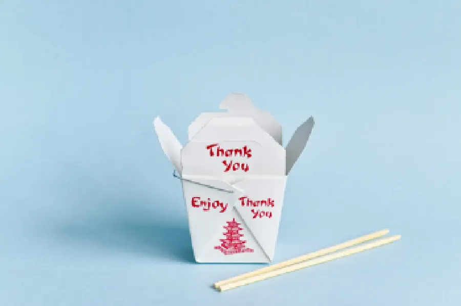 A white Chinese takeout box with red text reading "Thank You" and "Enjoy" sits open on a blue background next to a pair of wooden chopsticks.
