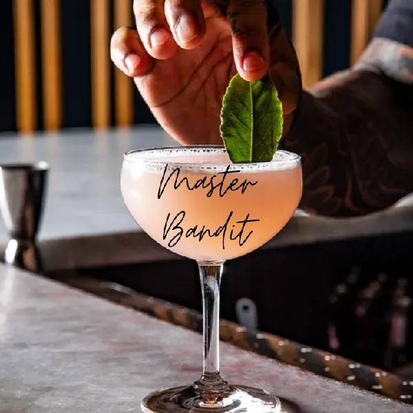 A hand garnishes a pink cocktail with a green leaf in a coupe glass on a bar counter. The words "Master Bandit" appear on the glass.