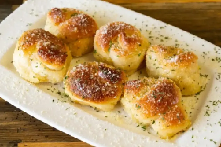 Six golden-brown garlic knots sprinkled with herbs and grated cheese are arranged on a white rectangular plate.