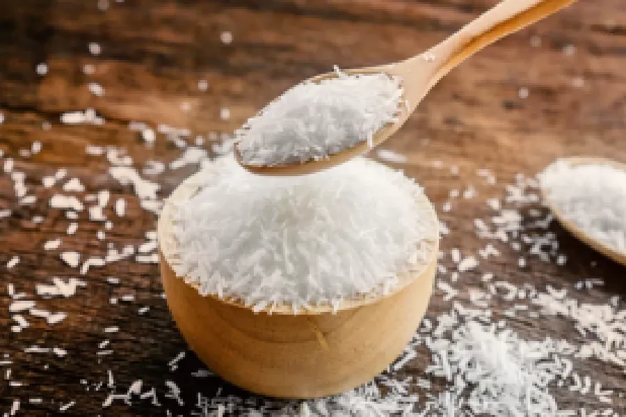 A wooden bowl filled with shredded coconut, with a wooden spoon holding more coconut above it, on a wooden surface.