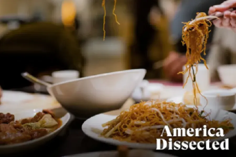 A hand lifts noodles with chopsticks over a table set with plates of food; text on image reads "America Dissected.