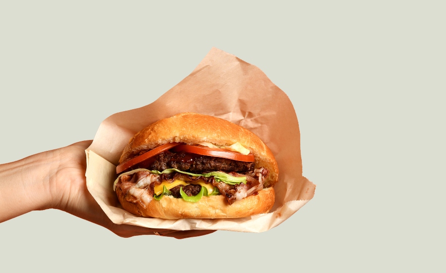 A hand holding a cheeseburger with lettuce, tomato, and melted cheese, wrapped in brown paper against a plain light background.