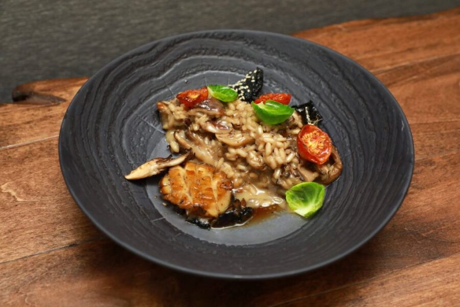 A black plate with mushroom risotto, roasted tomatoes, grilled mushrooms, brussels sprout leaves, and a light sauce, served on a wooden table.