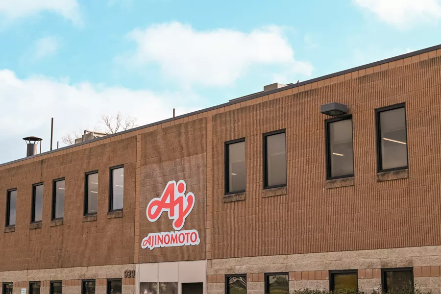 A brown brick building with the Ajinomoto company logo and name displayed near the entrance under a partly cloudy sky.