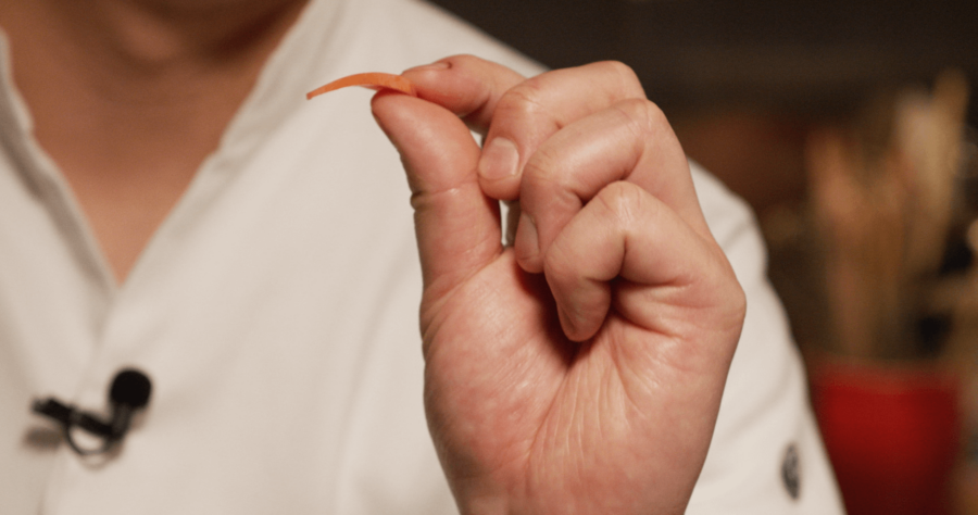 A person in a white shirt holds a thin orange slice between their thumb and forefinger.