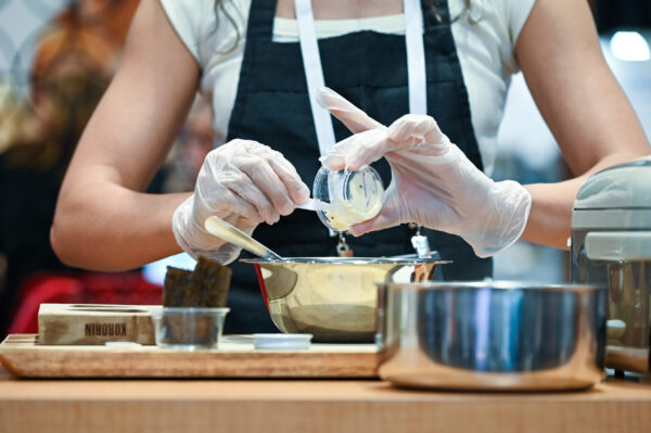 A person wearing gloves and an apron pours an ingredient from a small container into a mixing bowl on a kitchen counter with various utensils.