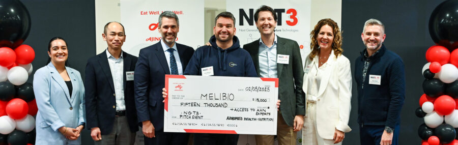 Seven people stand together, smiling, as one person holds a large check for $15,000 made out to MeliBio at an event with branded banners and balloons in the background.