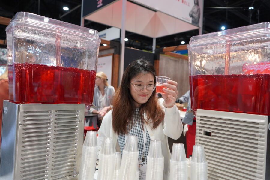 A woman stands between two large dispensers of red beverage, holding a small sample cup, with stacks of clear plastic cups in front of her at an indoor event.