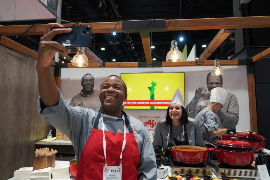A chef in a red apron takes a selfie while another chef in a white hat smiles beside him at a food event booth with pots and a promotional display in the background.