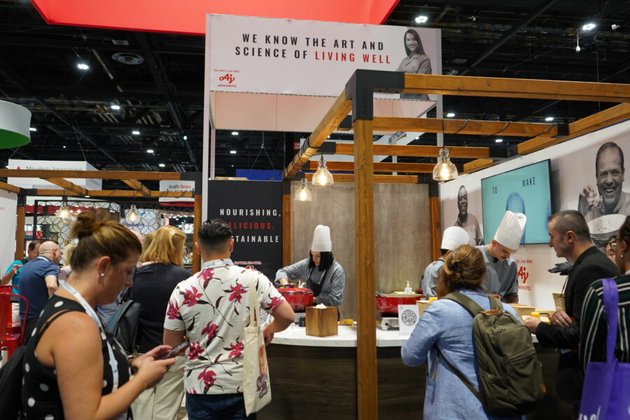People gather around a food booth at an exhibition, where chefs in white hats prepare dishes under a sign reading "We Know the Art and Science of Living Well.