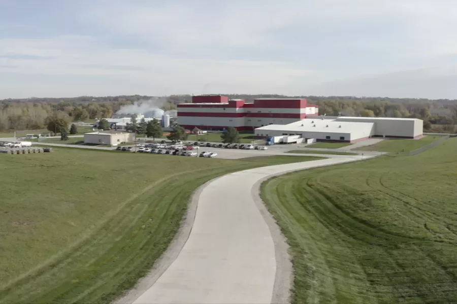 A large industrial facility with red and white buildings, surrounded by green fields, parking lots, and a winding paved road under a partly cloudy sky.