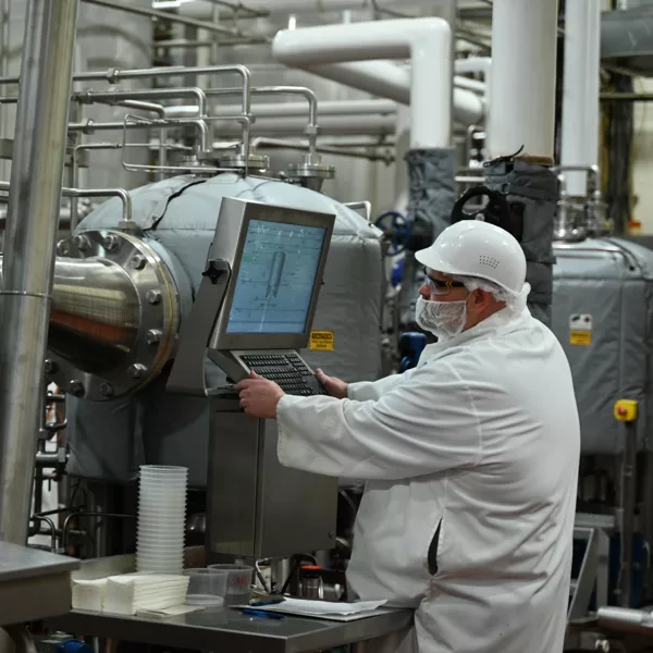 A worker in protective clothing and a hard hat operates a touchscreen control panel in an industrial facility with metal pipes and equipment.