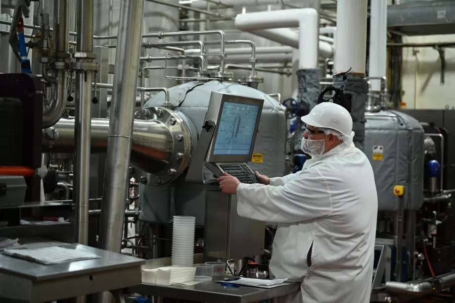 A worker in protective clothing and a hairnet operates a computer terminal in an industrial processing facility with pipes and machinery in the background.