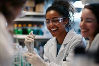Three scientists wearing lab coats and safety glasses smile and work together in a laboratory, handling lab equipment and bottles.