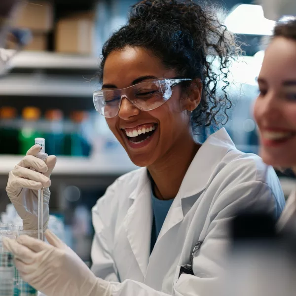 A scientist wearing safety goggles and gloves smiles while handling a pipette in a laboratory setting, with another person beside her.