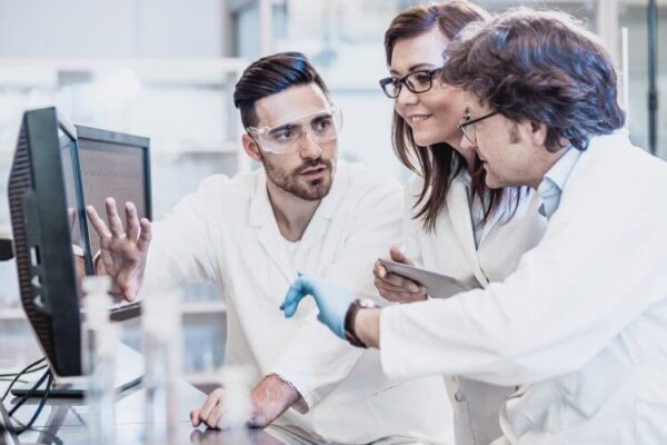 Three scientists in lab coats review data on a computer screen in a laboratory, with one person holding a tablet and another wearing safety goggles and gloves.