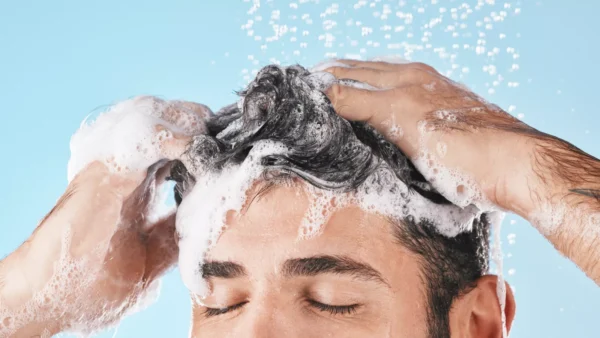 A person with closed eyes is washing their hair with shampoo, creating lather and bubbles under running water against a blue background.