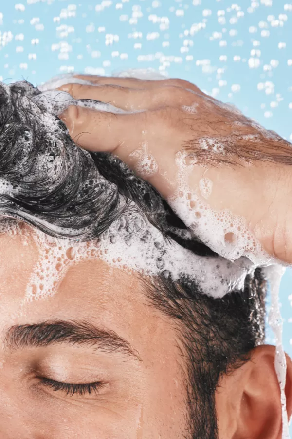 A person washing their hair with shampoo, creating lather and bubbles, with water droplets and a blue background visible.