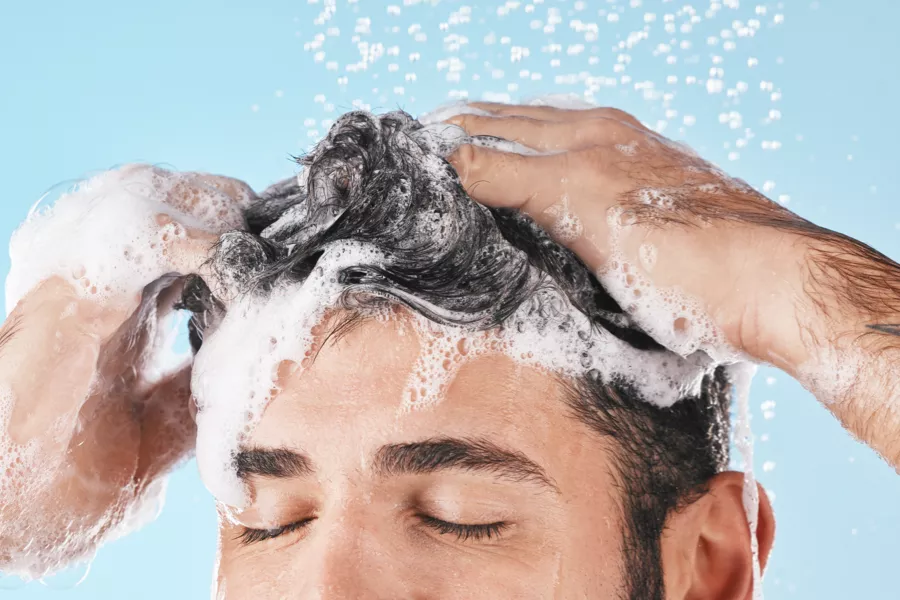 A person with closed eyes washing their hair, lathered with shampoo and water, against a blue background.