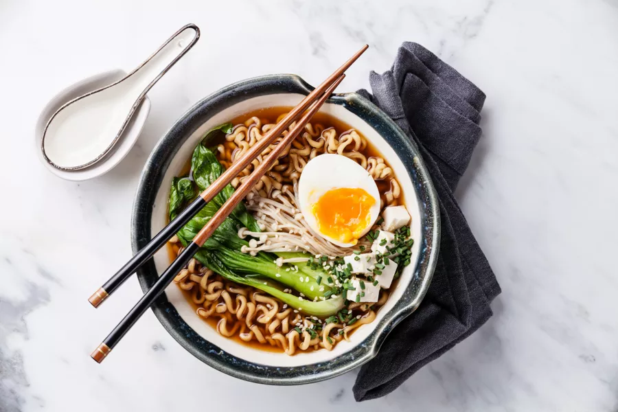 A bowl of ramen with noodles, bok choy, tofu, enoki mushrooms, a soft-boiled egg, and chopped chives, served with chopsticks on a marble surface.