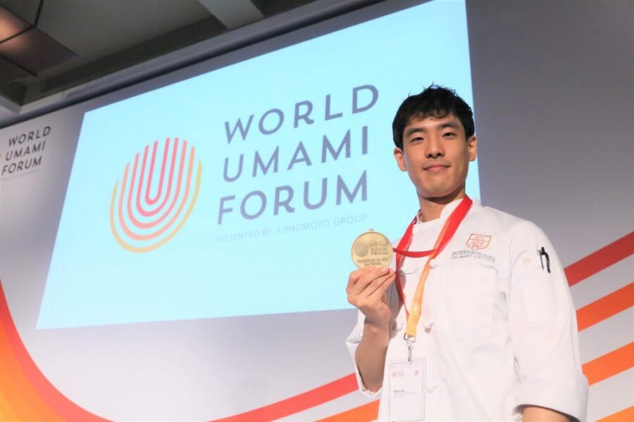 A chef in a white uniform holds a gold medal while standing in front of a large "World Umami Forum" sign on stage.