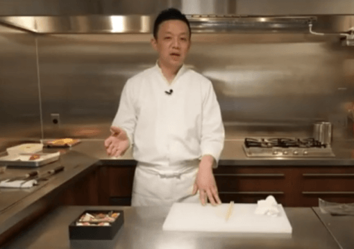 A chef in a white uniform stands in a stainless steel kitchen behind a counter with a cutting board, chopsticks, and a box of food.