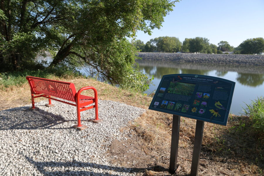 A red bench and informational sign about pollinators are positioned on gravel beside a calm river, with trees and greenery in the background.