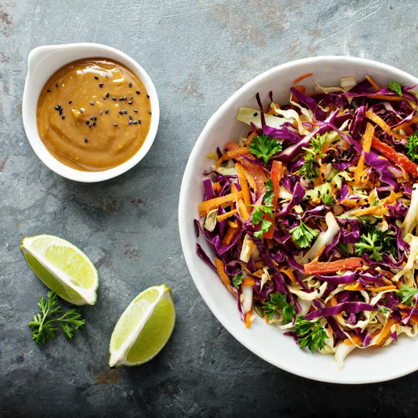 A bowl of colorful cabbage slaw with carrots and parsley next to lime wedges and a small dish of creamy dressing sprinkled with black sesame seeds.