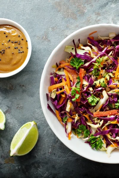 A bowl of shredded red and green cabbage salad with carrots and parsley, served with lime wedges and a side of sesame ginger dressing.