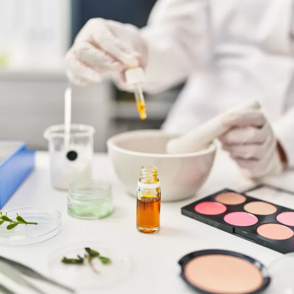 Person in lab coat and gloves uses dropper with amber liquid, surrounded by laboratory glassware, a mortar and pestle, plant samples, and a makeup palette on the table.
