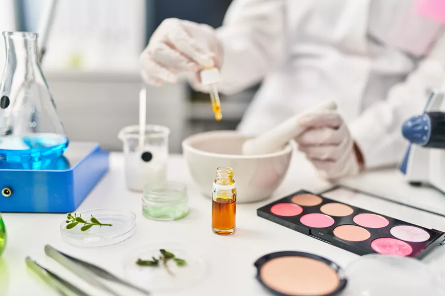 A person in gloves mixes substances in a laboratory with scientific glassware, a makeup palette, plant samples, and a dropper bottle on the table.