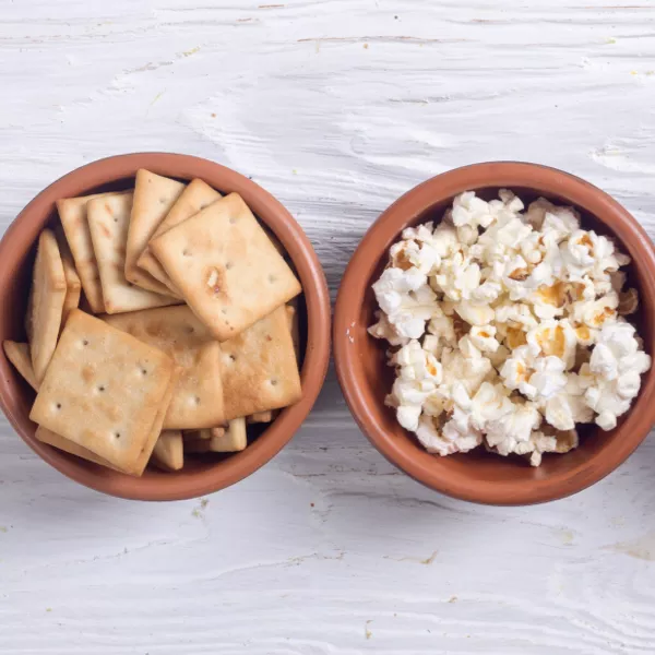 Two brown bowls on a white surface, one filled with square crackers and the other with popped popcorn.